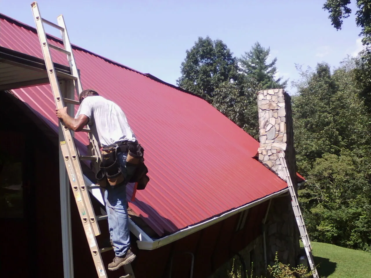 Roofer on a ladder beside a red metal roof and stone chimney — Soffit & Fascia Repair by Roofing Brothers in Stites